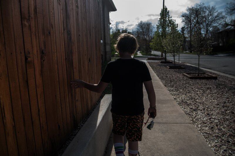 A 13-year-old boy walks on a sidewalk next to a wooden fence. You can’t see his face. The photo is taken from the back. He is wearing a T-shirt.