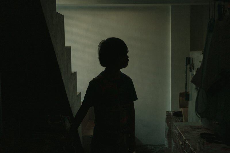 A boy stands silhouetted in a dark hallway next to a counter and a set of stairs.