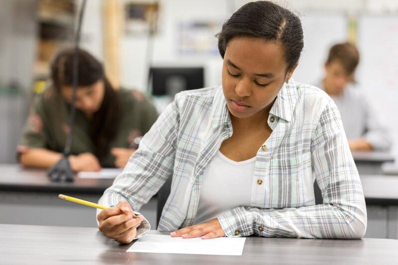 A high school student sits at a black table taking an exam with a yellow No. 2 pencil. Two students sit at black tables behind her taking an exam.