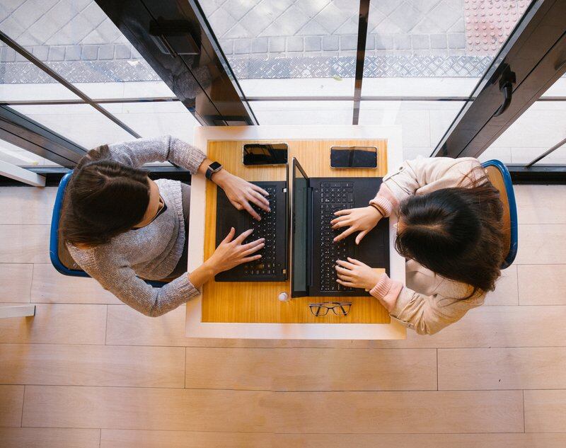 Two young women work at their laptops in front of a window.