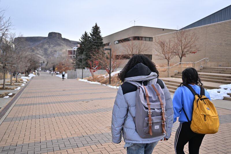 Two students with backpacks on walk on on a college campus with a mountain in the background.