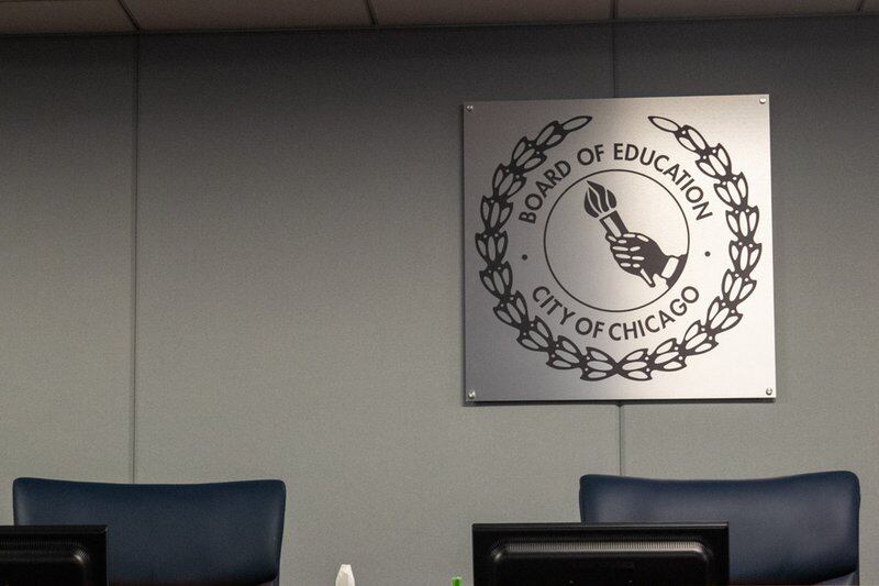 A photo of two empty seats for Chicago’s school board members on shared dais. In front of the seats is the table part of the dais. Behind the seats is a gray, matte, and square sign. In the middle of the sign is a circle with a hand holding a torch inside. Above the circle is the words “Board of Education,” curved around the circle. Below is the circle is the words “City of Chicago,” also curved. Wrapping around the words in a half circle is a laurel.