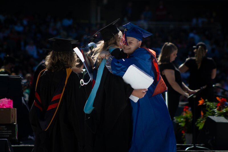 A young woman and educator embrace during a graduation ceremony, both wearing full graduation regalia.