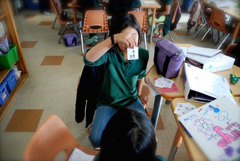 A student wearing a green polo shirt holds up a math flashcard showing the problem 6 x 6 for a classmate. The student is seated and their face is obscured by the card.