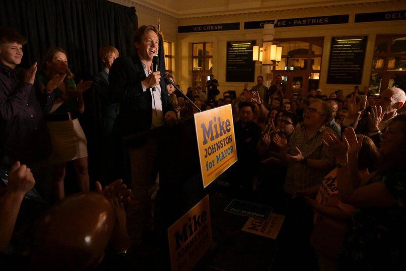 A man, Mike Johnston, stands at a darkened room from a podium with a  “Mike Johnston for mayor” sign on front of it and addresses a crowd of people.