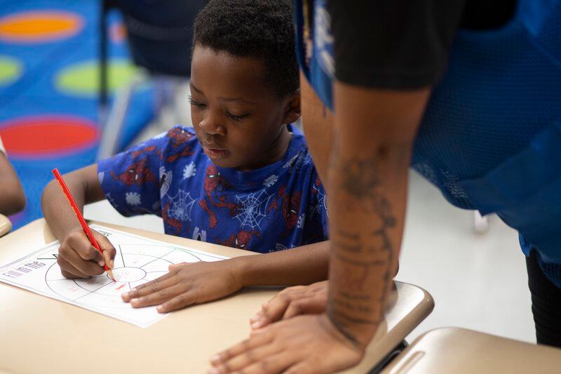 A boy wearing a blue shirt sits at a desk as a teacher’s aide helps him with his math work.