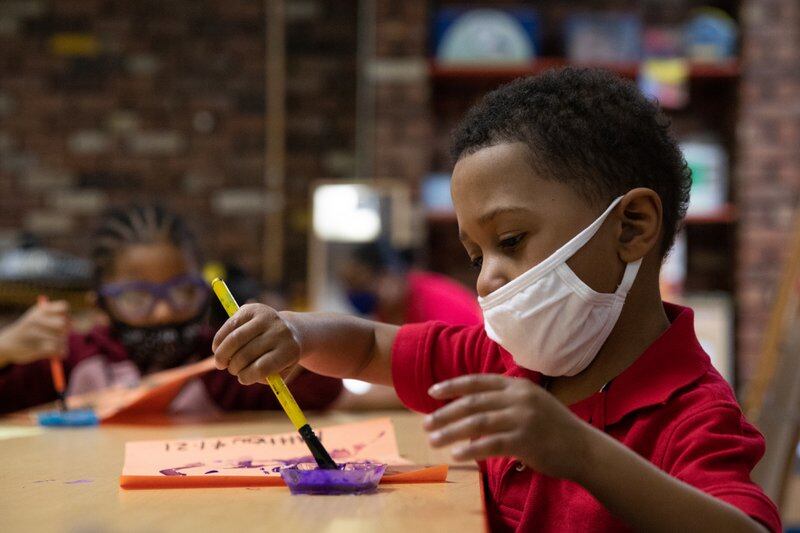 A young boy sitting in front of a piece of paper dips his brush in paint while at a preschool class.