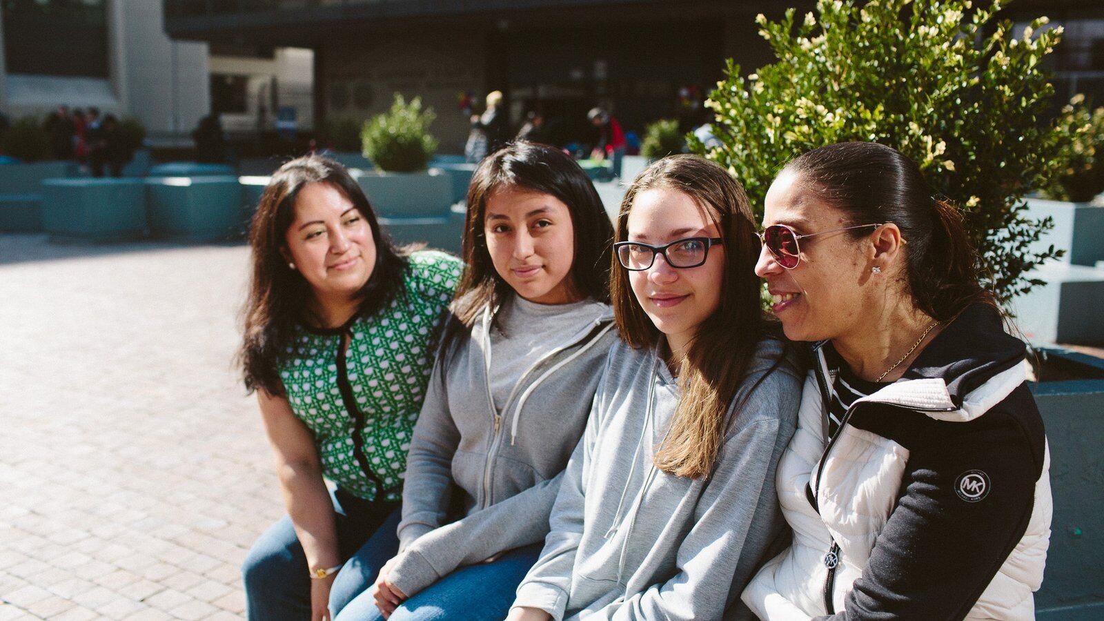 From left, Lordes Lliguichiuzhca, her daughter Elizabeth Cuzco, 13, Sonia Bucur, 13, and her mother Dina Bucur, sit in front of Martin Luther King High School after attending the Round 2 high school fair.