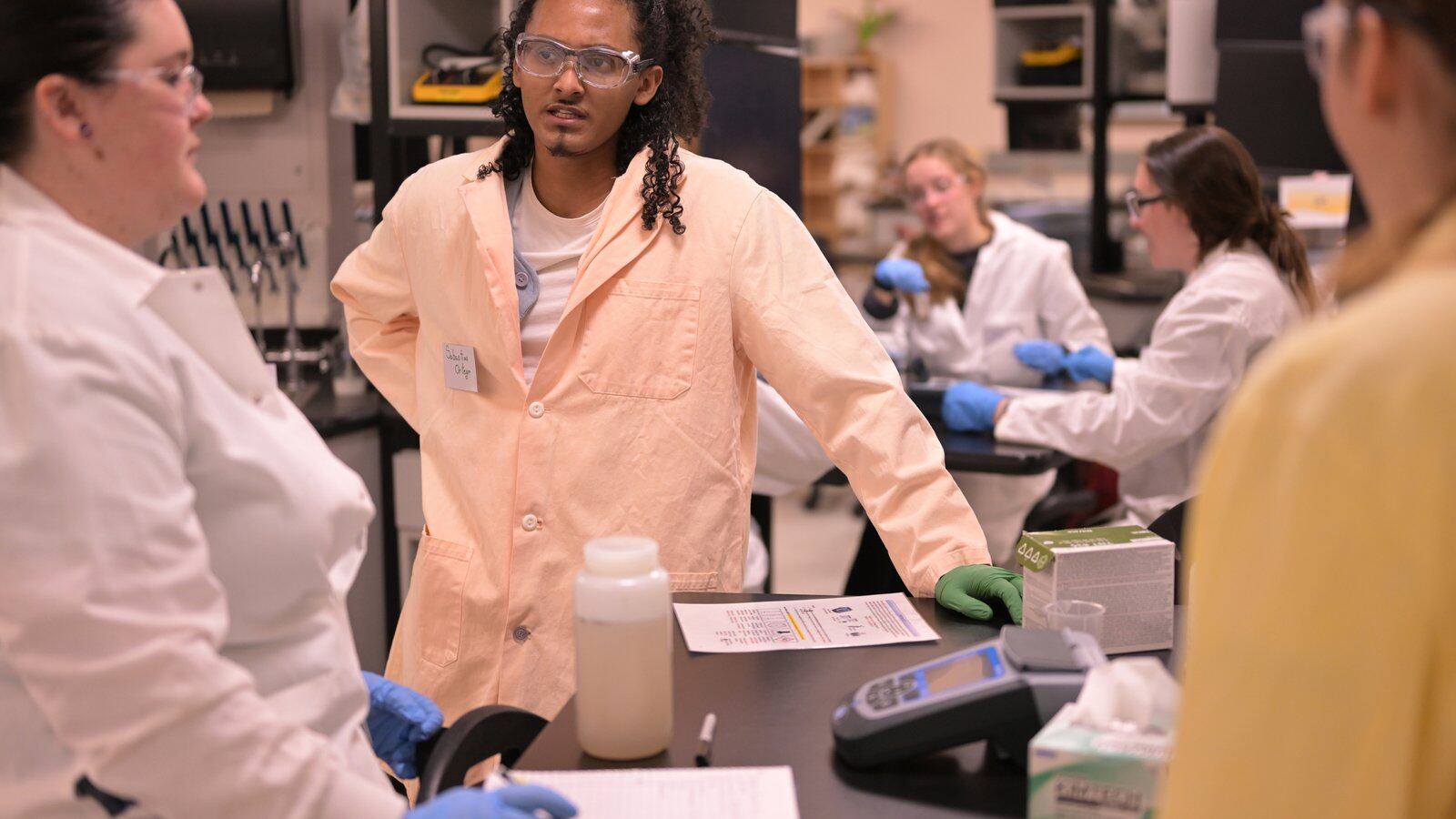 Students wearing lab coats and safety equipment work in a college laboratory.