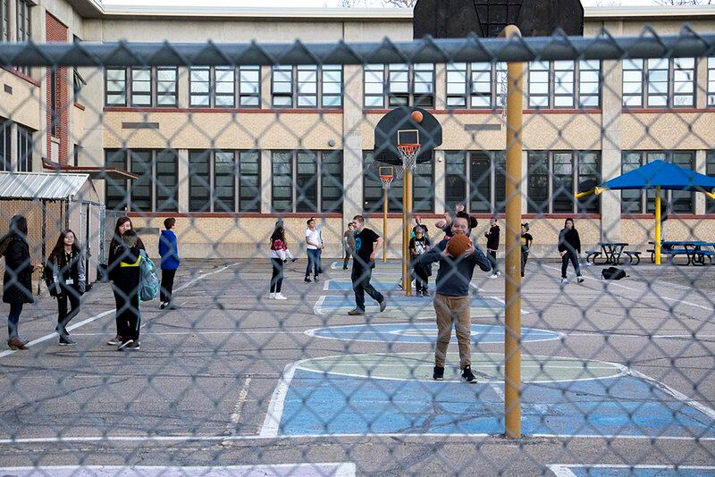 Middle school students stand on the blacktop outside a brick school building. They are seen through a chain link fence.