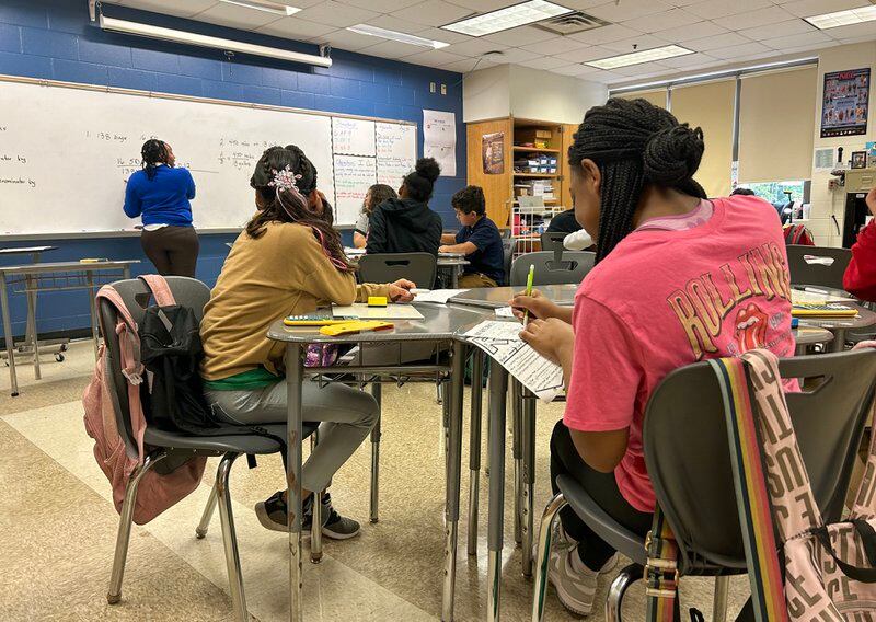A student wearing a pink shirt sits at a table with other students in a classroom. In the background, a teacher in a blue top writes on the whiteboard.
