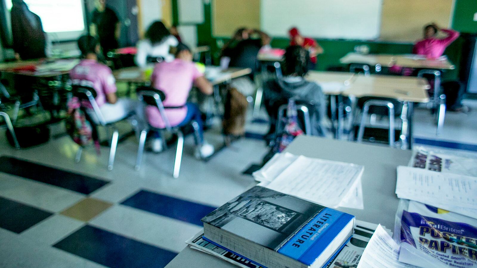 Textbooks are in the foreground with students seated at desks in the background.