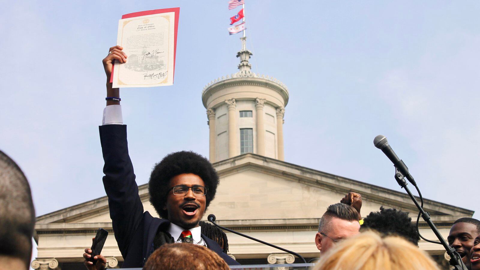 A man with glasses and an Afro style haircut stands with his hand in the air holding a piece of paper.