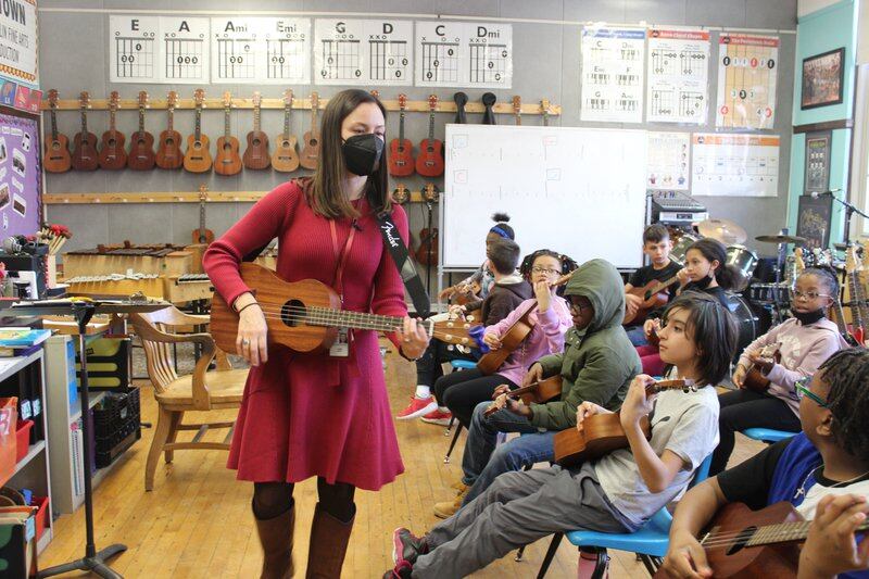 A teacher in a red dress demonstrates how to play a ukulele for her fourth grade classroom.