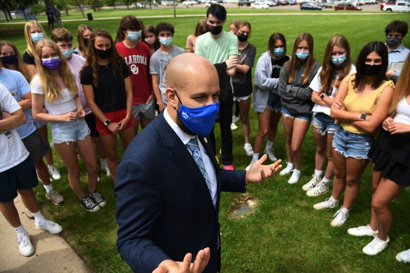 Alex Marrero, standing outside on grass and wearing a mask, is surrounded by masked high school students listening to him talk.