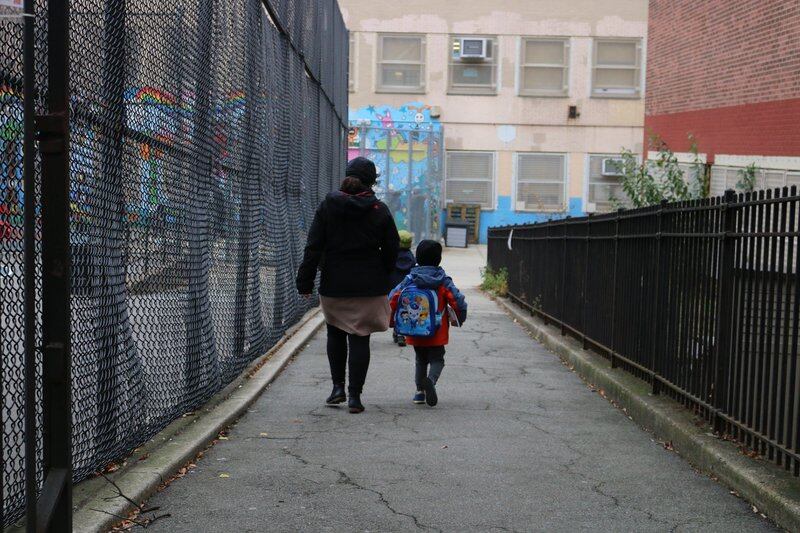 A parent and a child walk toward an elementary school, with fencing on either side. Their backs are turned to the camera.