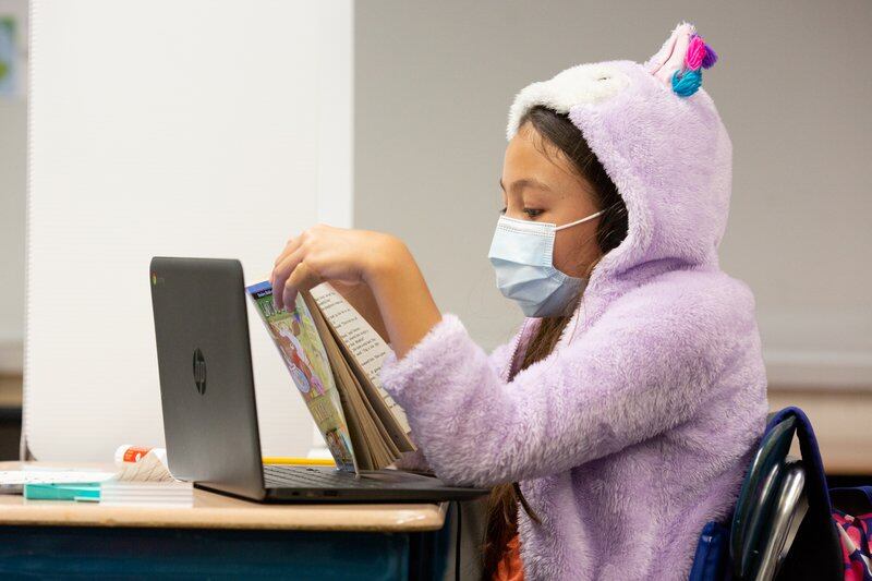Student wearing a mask and lavender hooded sweatshirt with ears is reading a book in class.