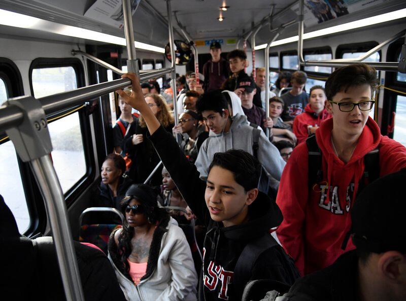 A city bus is packed with teenagers, many of them wearing hoodies, hanging from the hand rails.