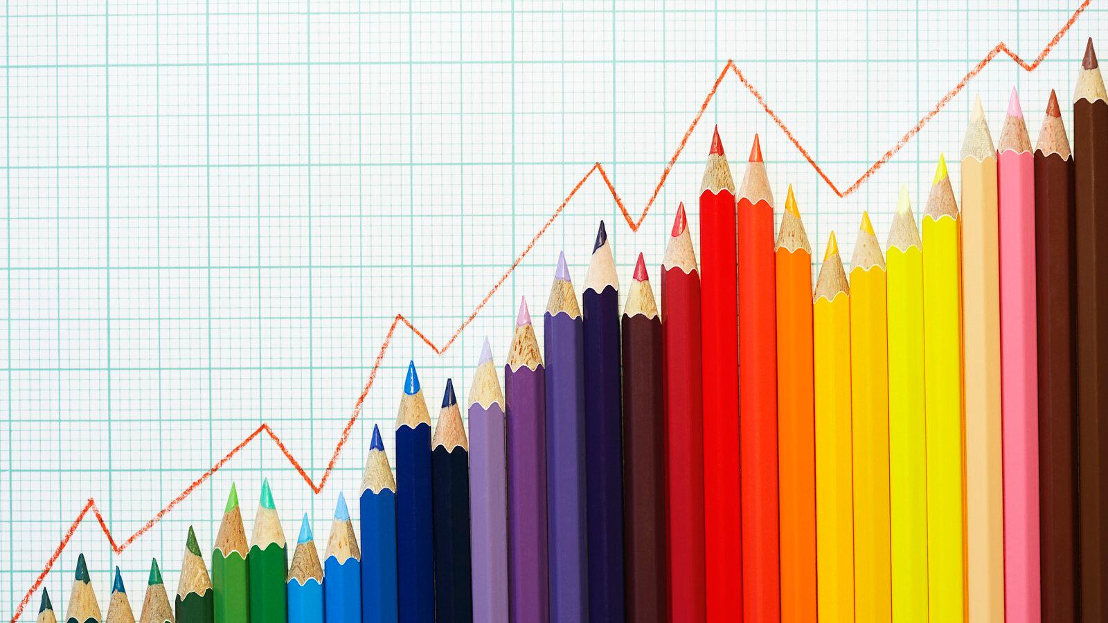Colored pencils are lined up in the shape of a graph with a read line following the top of the pencil tips with white and blue graph paper as the background.