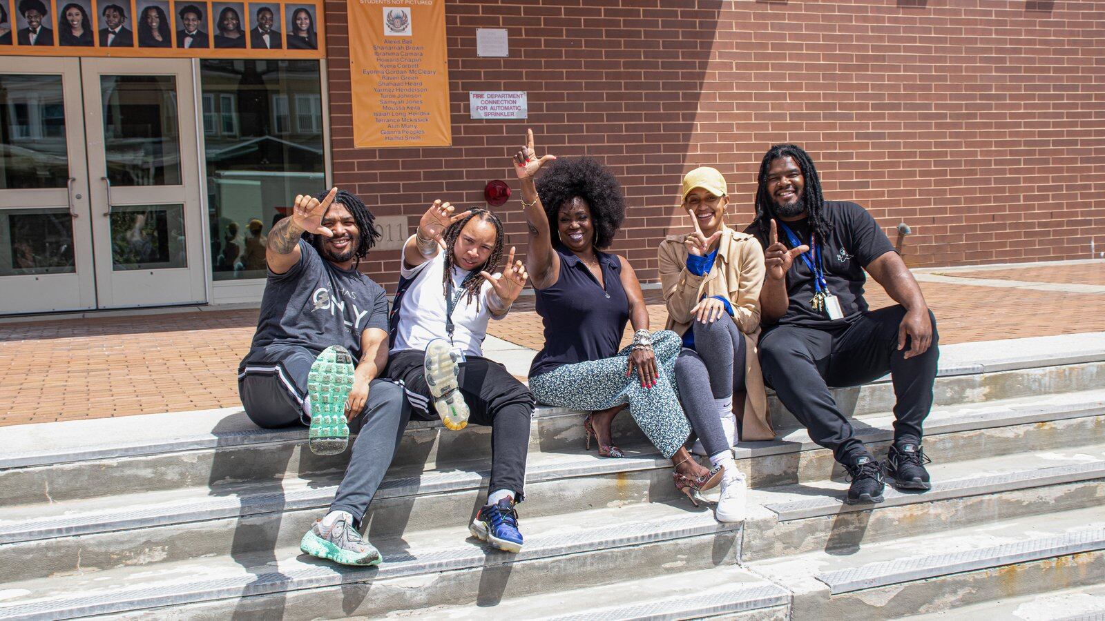 Five people pose and gesture during a portrait on the steps of West Philadelphia High School.