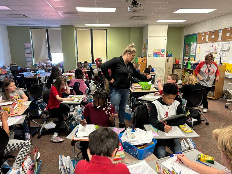 A woman in jeans and a black top with blonde hair stands in the middle of a classroom where several children are sitting at desks.