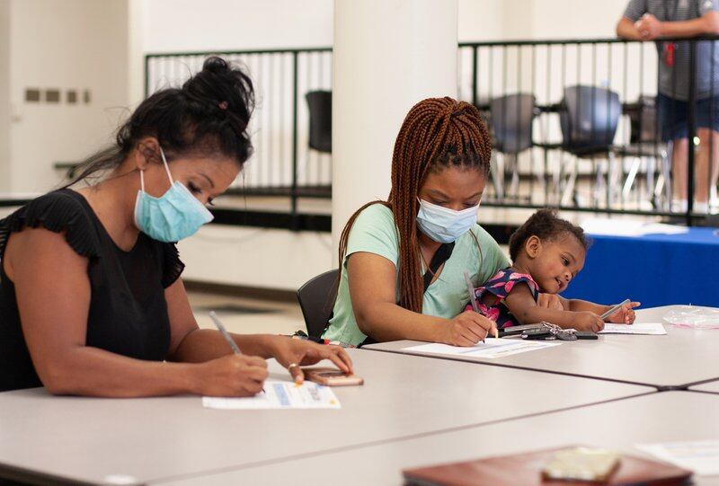 A woman in a black shirt wearing a blue face mask with her hair in a bun and a woman in a teal shirt with a blue face mask holding a distracted young child on her lap sit at a table and write on pieces of paper.