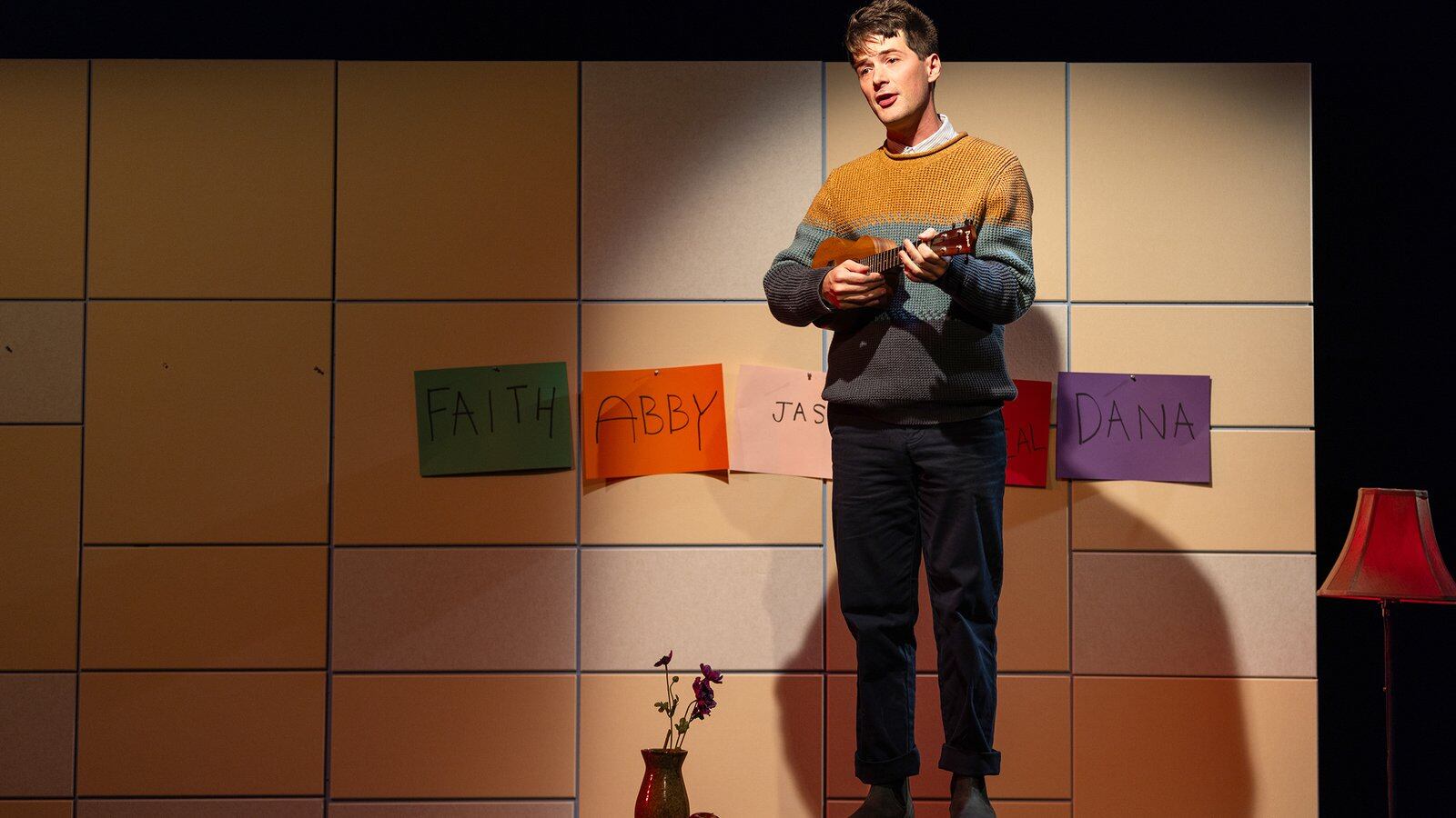 A person with short hair and wearing a multicolored sweater performs on a stage playing a ukulele.