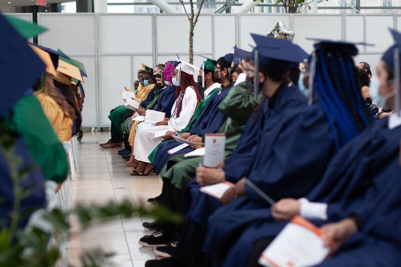 Rows of graduating students wearing blue, yellow, white and green graduation gowns and caps listen to a speaker.