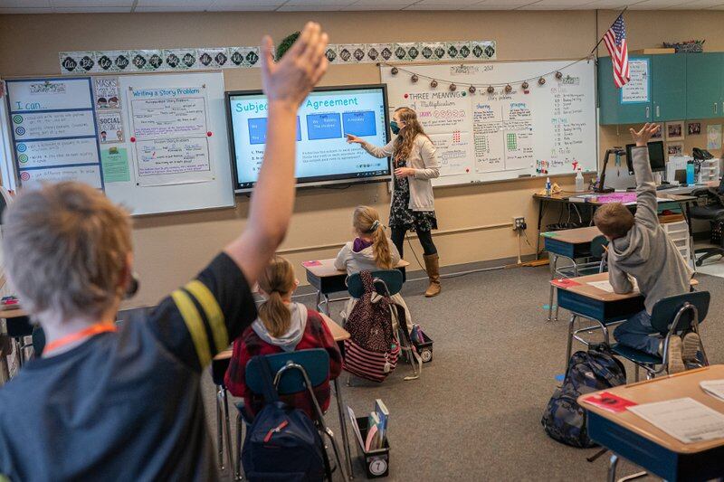 A young student raises his hand in the foreground as his classmates listen to their teacher at their desks. The teacher is pointing at a smartboard at the front of the classroom.
