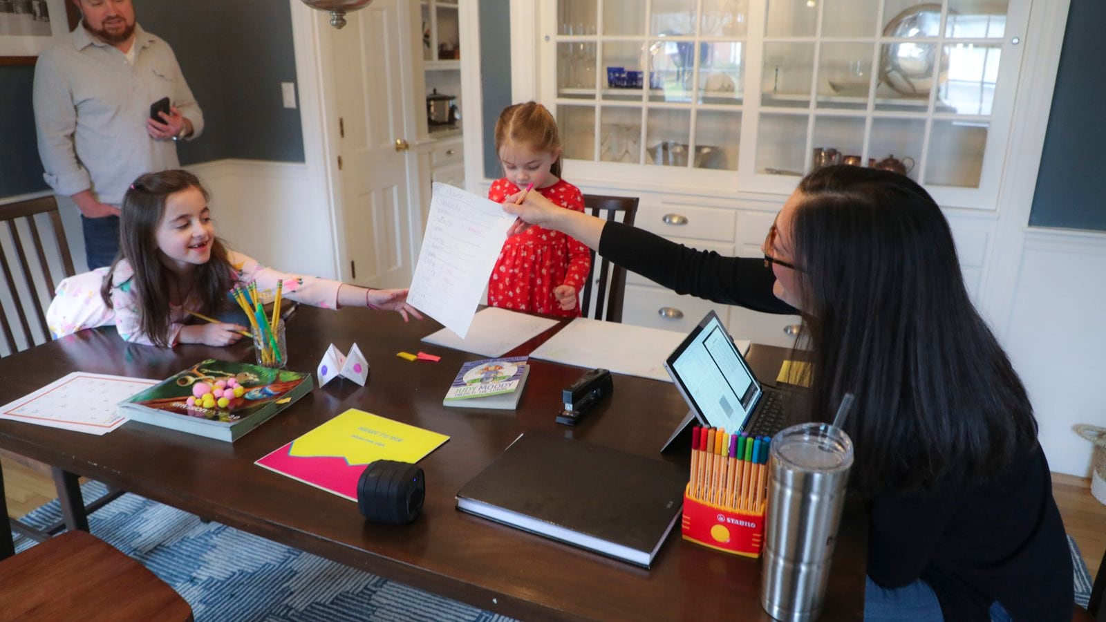 Sarah Yunits checks her daugher Ada's homework while Cora waits her turn as dad Conor Yunits is on a work conference call at their home in Brockton on March 19, 2020. Because of the coronavirus, the Yunits are able to perform their jobs at home and are home schooling their two children.