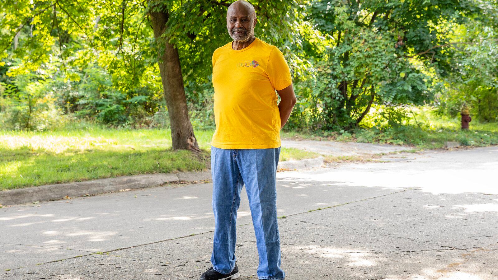 An adult man wearing a yellow shirt and blue jeans poses for a portrait in the middle of a street with green trees and grass in the background.