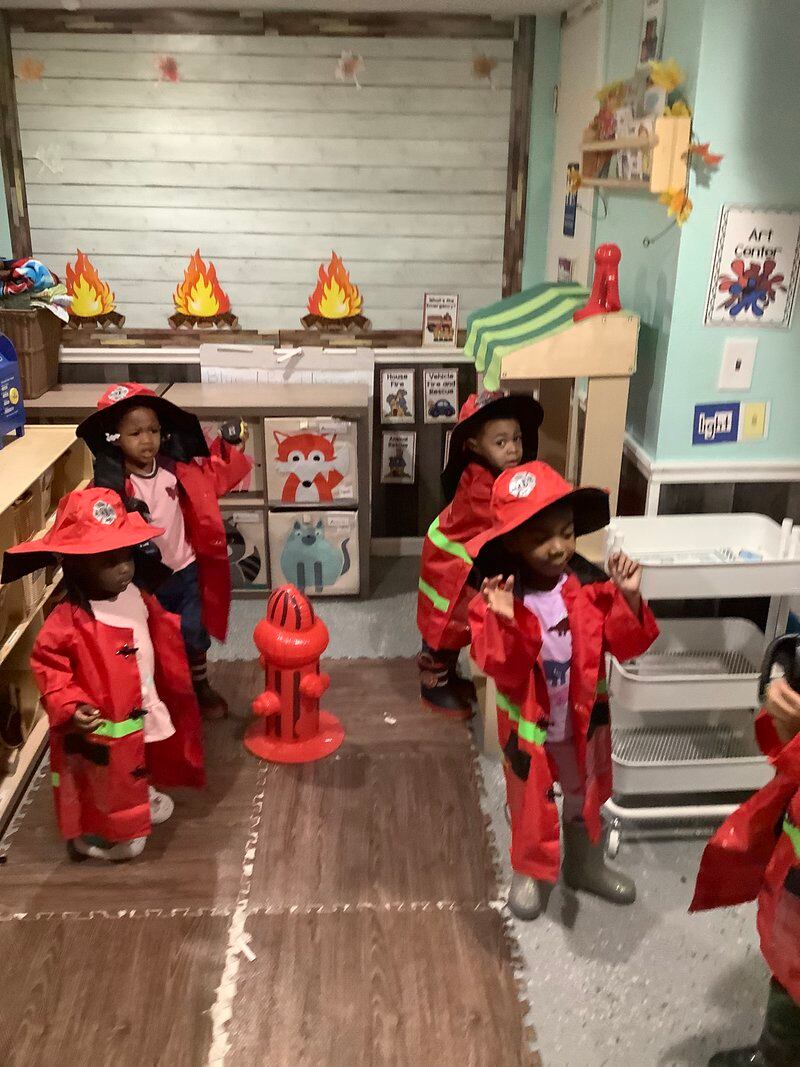 Children wear pretend red fire outfits and helmets next to a pretend red fire hydrant in a preschool classroom