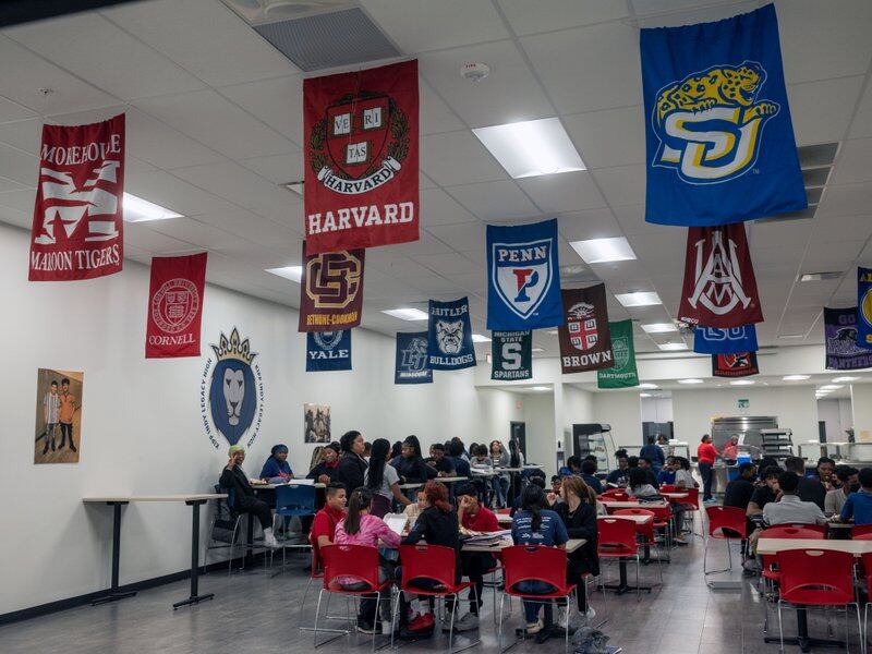 Flags of various colleges hang from the ceiling of a high school cafeteria as students sit at their tables.