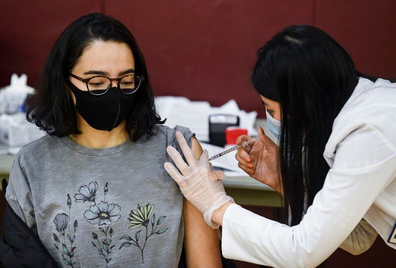 A young woman wearing glasses, a black mask and a grey t-shirt with flowers receives a vaccine from a woman wearing a blue surgical mask, gloves and white coat.