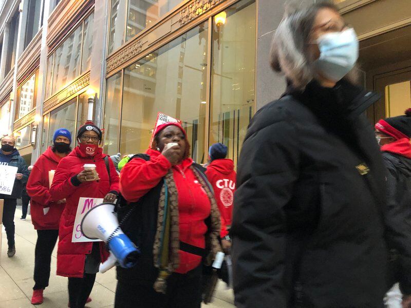 A group of people wearing red carrying posters and a megaphone march on a downtown sidewalk.