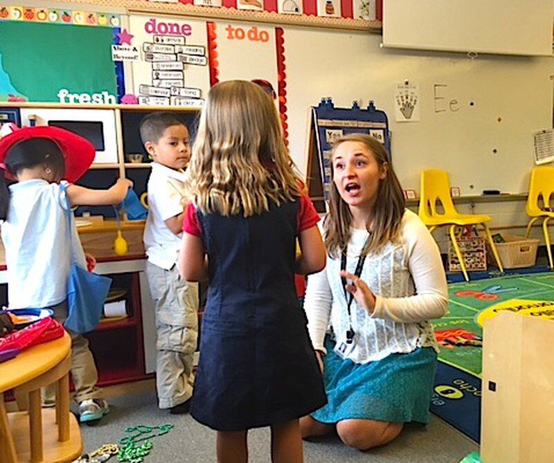 A preschool teacher sitting on the floor talks with one of her students, who is facing her. 
