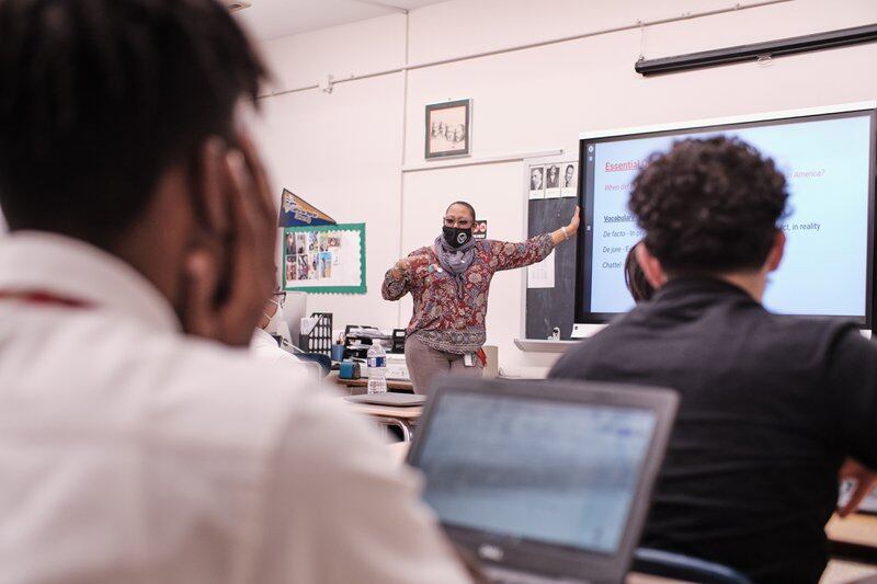 A teacher wearing a black face mask and colorful flowered shirt points to a whiteboard while two students look on from their desks with their laptops open.