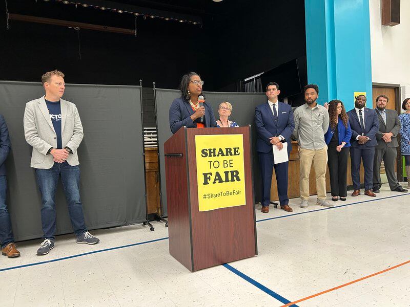 A woman holding a microphone stands behind a podium with a yellow sign on it that reads, “share to be fair.”