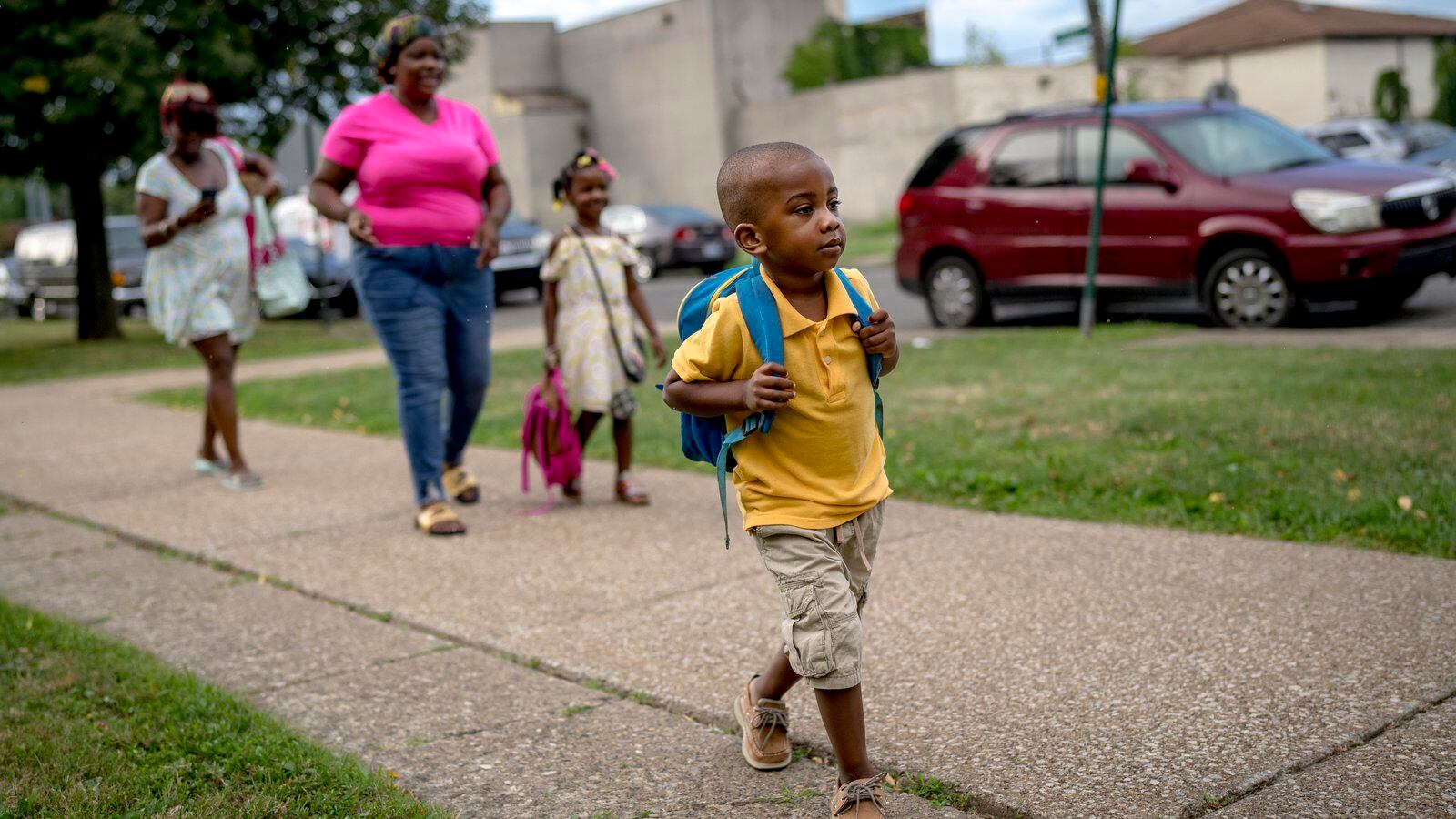 A child wearing a backpack walks toward a school building. Other children and parents can be seen behind him.