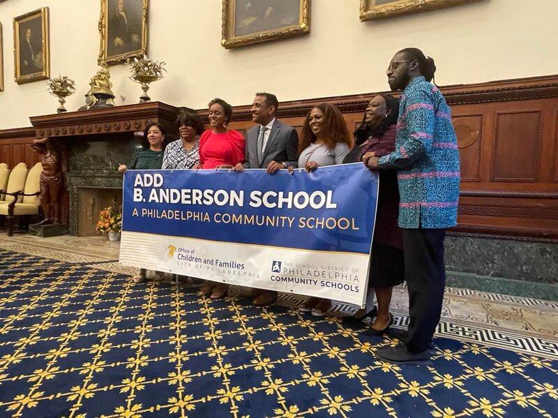 Seven people hold up a blue and white banner with the name “Add B. Anderson School” written in white.