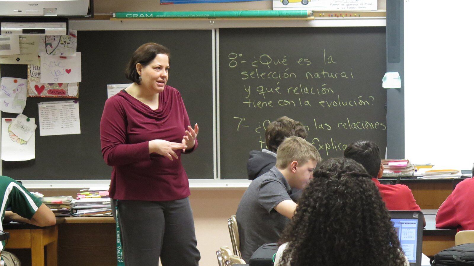A woman in a maroon blouse instructs her class, standing in between the desks of her students