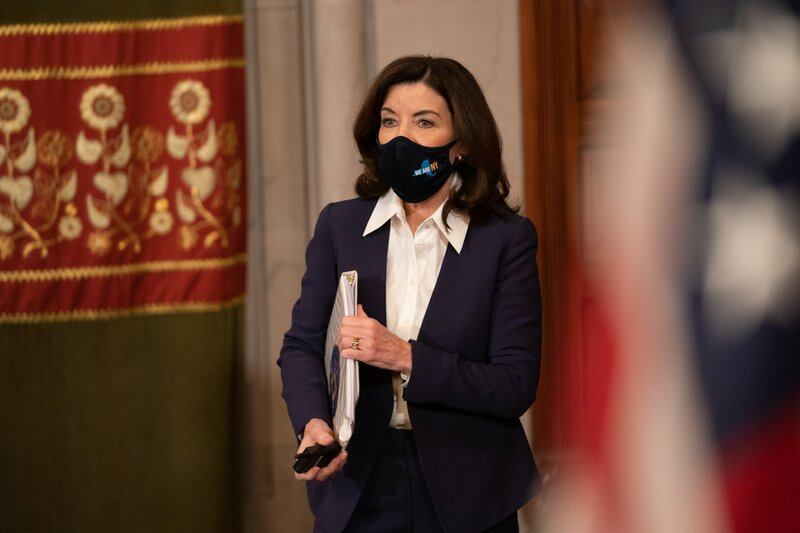 Gov. Kathy Hochul, wearing a mask and a dark suit and carrying a briefcase, gives a presentation in a room with curtains and a flag.