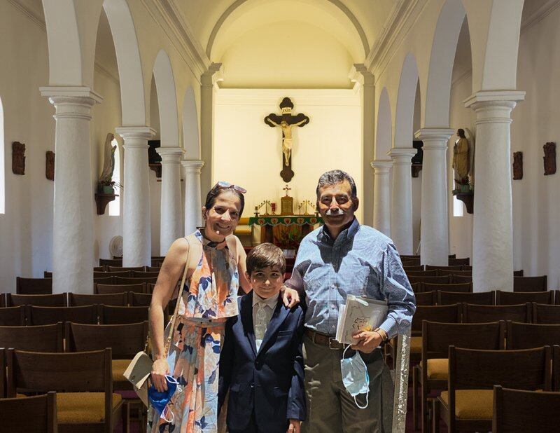 A mother and father stand in a church with a 10-year-old boy between them. They all smile at the camera. A crucifix can be seen in the background.