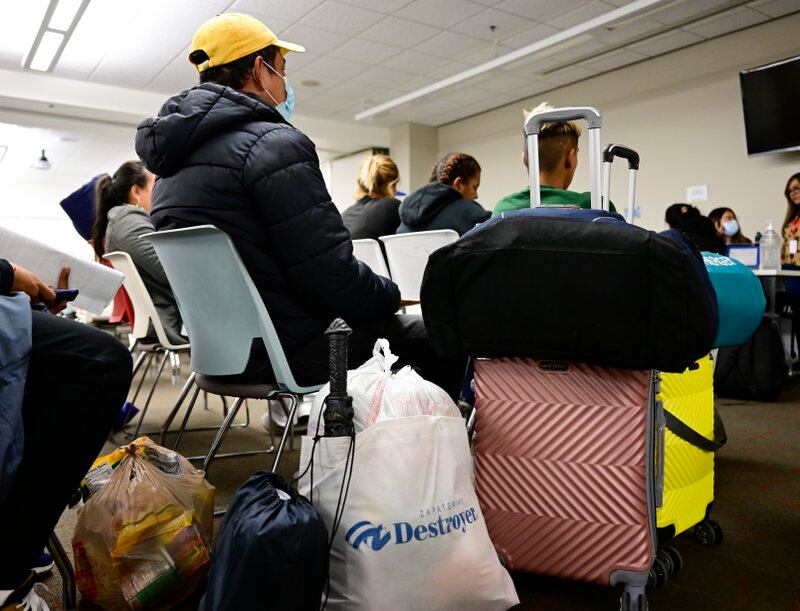 Bags of different sizes and a pink suitcase sit in front of a person sitting in a chair wearing a black jacket and a yellow cap. People sit in chairs waiting in the background.