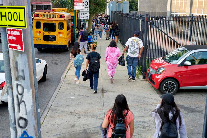 Students, seen from behind, walk on the side walk next to a yellow school bus. 