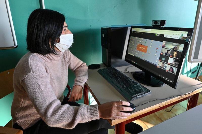 A masked female student sits at a desk and looks at a screen showing a virtual meeting.