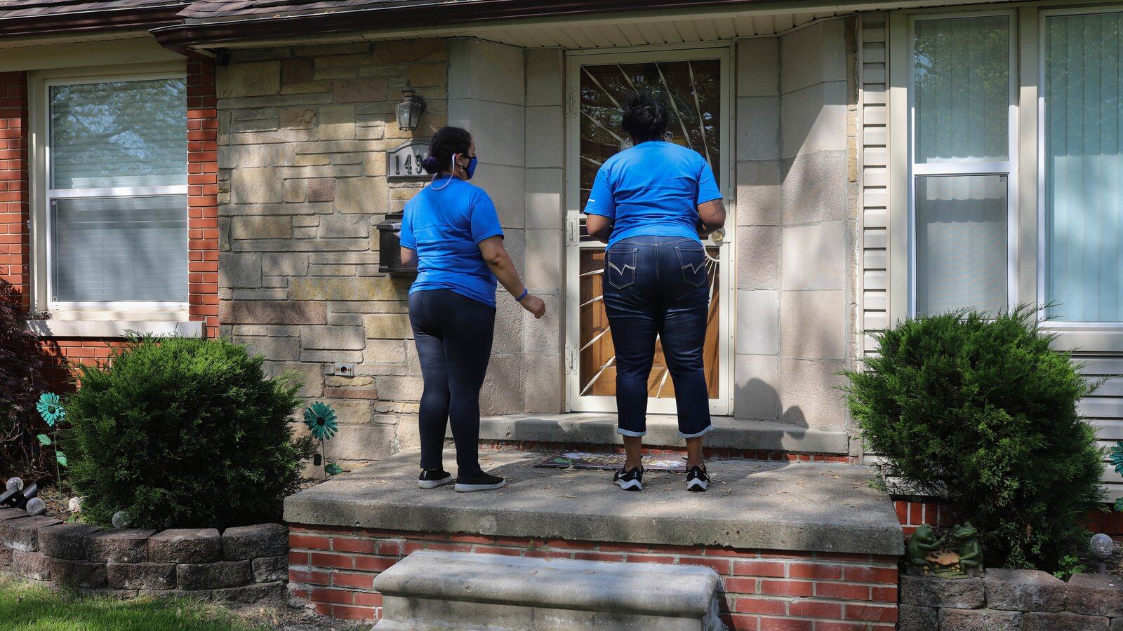 Two women, wearing blue shirts and jeans, stand at the front door of a house in Detroit, Mich. that is patterned from the sun and shadows.