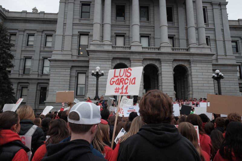 Students dressed in red gather in front of the Colorado State Capitol. There’s a large crowd of young people. Classical columns rise before them. One sign held aloft reads “Luis Garcia #11.”