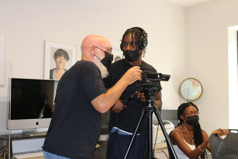 A man in a mask leans over a video camera while a student stands beside him.