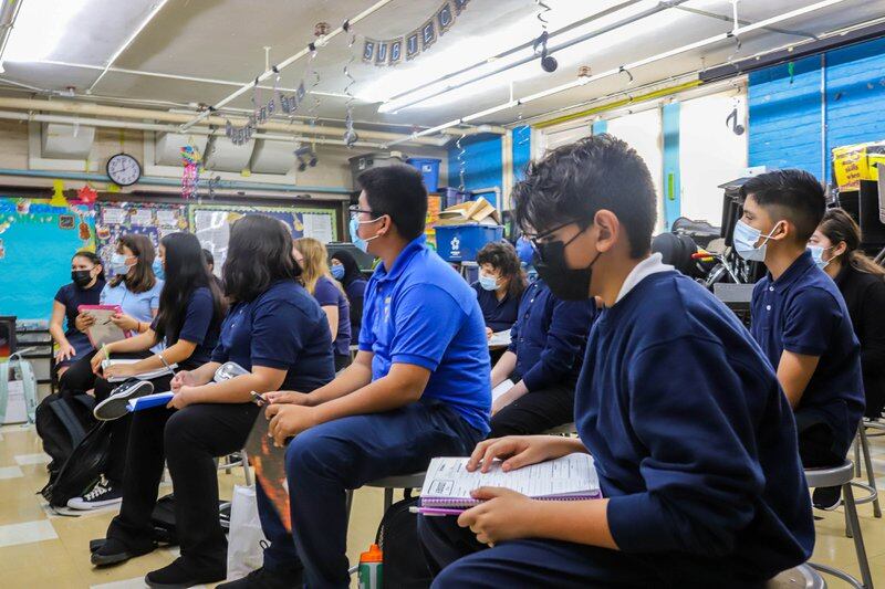 Masked students sit in rows in a classroom, all wearing blue shirts and sweaters.